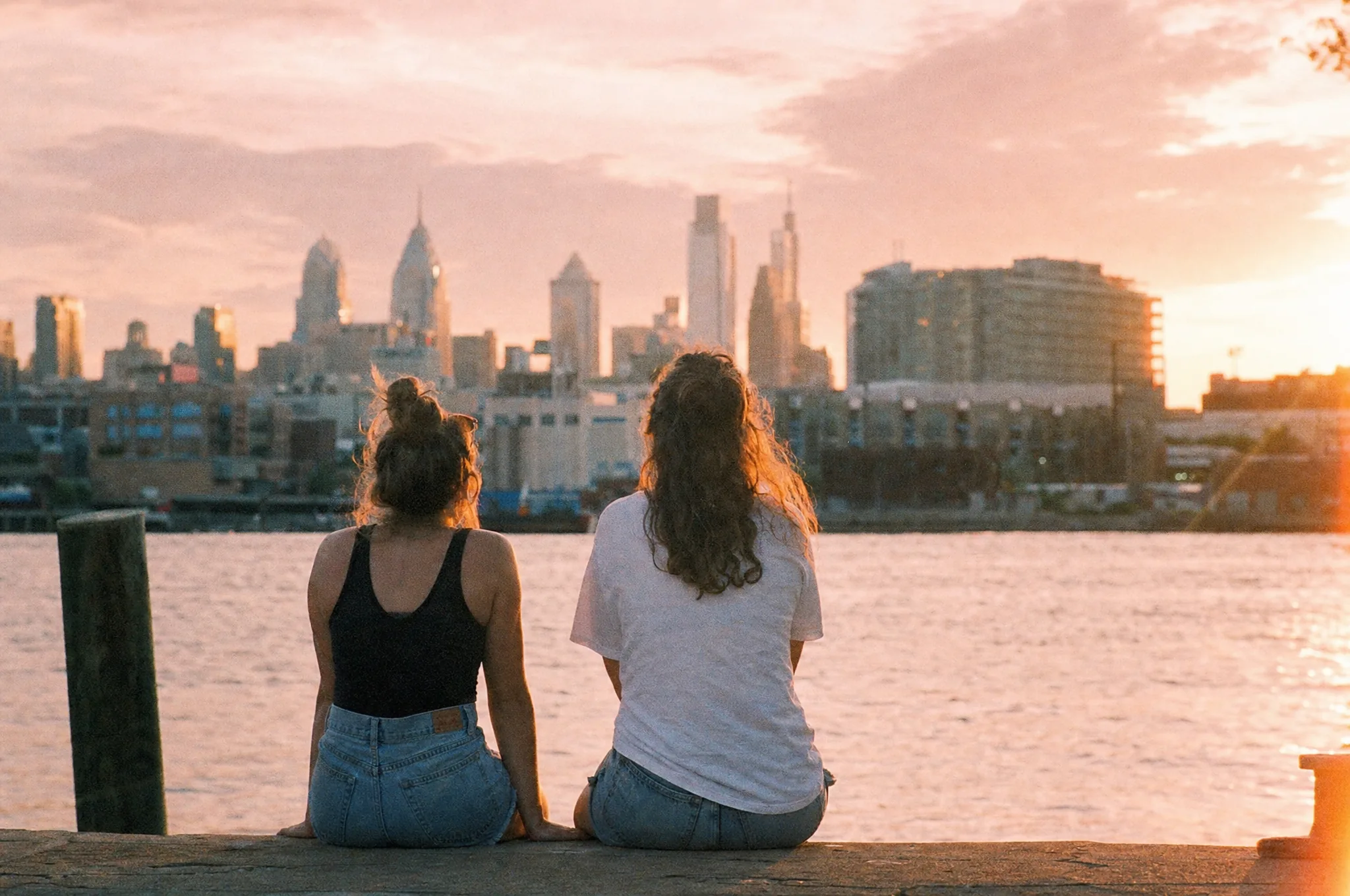Two friends sitting on a river pier at sunset with the Philadelphia skyline behind them